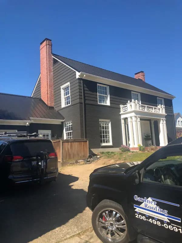 Black modern house with a white porch and chimney, featuring a Gardiner Roofing vehicle nearby.