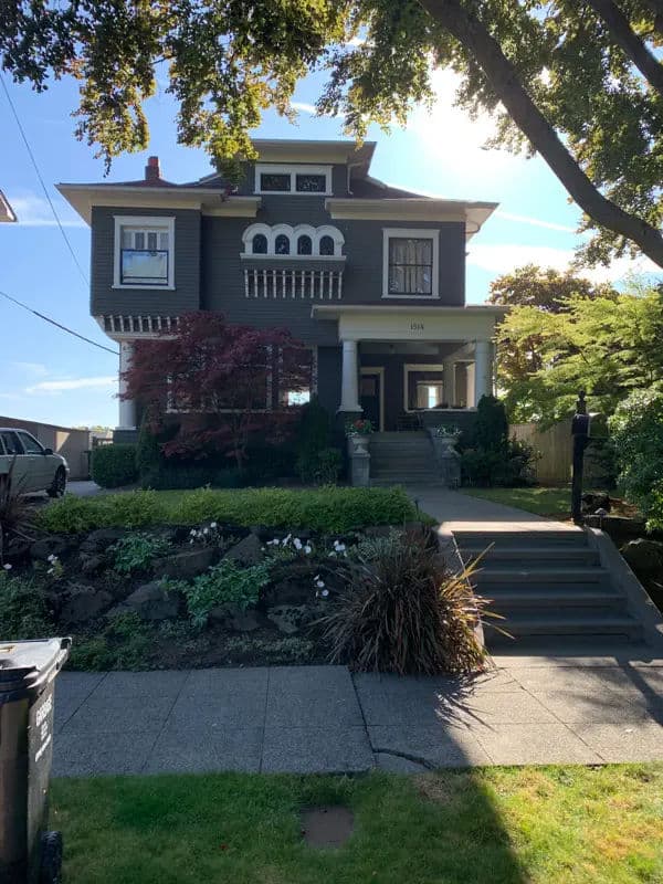 Historic gray house with porch, lush garden, and unique architectural details in a sunny setting.