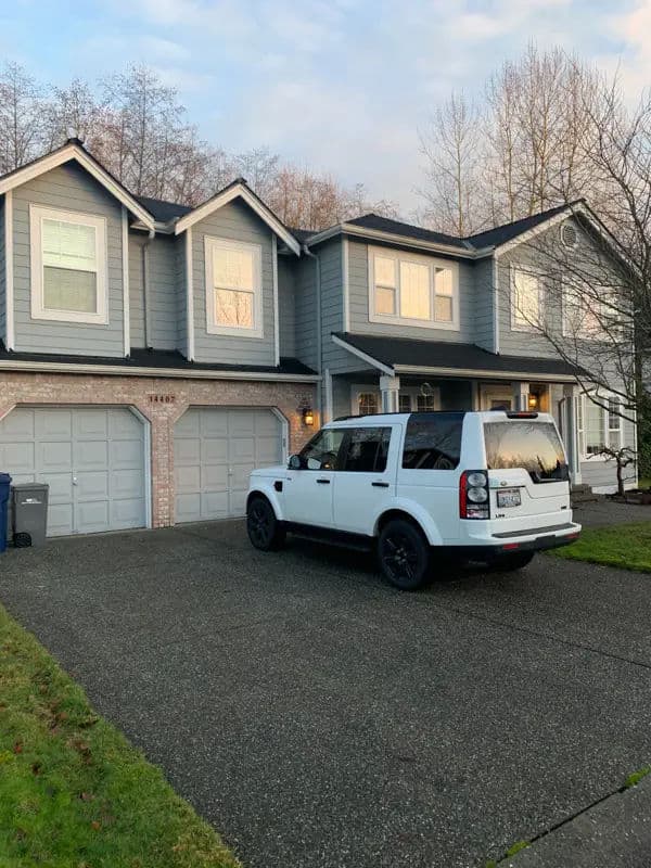White SUV parked in front of a gray two-story house with a landscaped yard.
